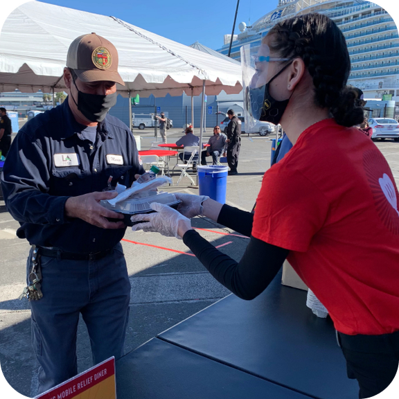 A woman with a face mask and gloves is giving a meal to a man wearing a baseball cap and a face mask